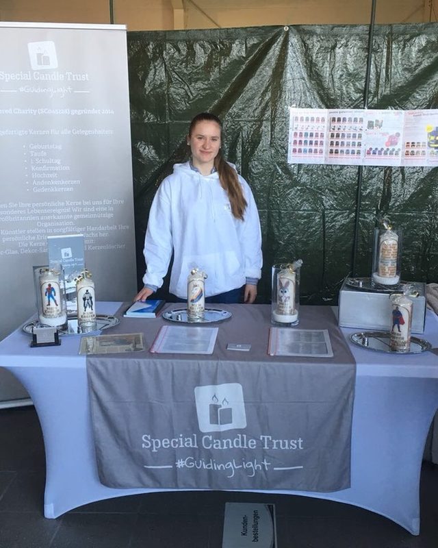 A young volunteer at a Special Candle Trust stand with a German-language banner and candles on display